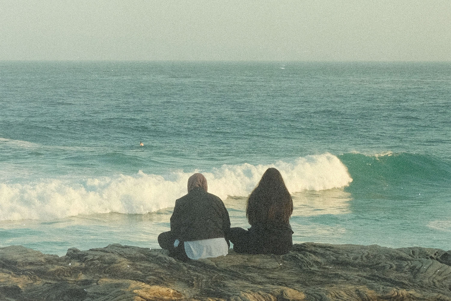 Two people sitting on rocks overlooking ocean waves rolling toward the shore