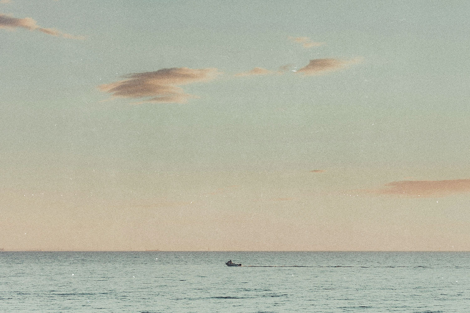 Small boat moving across a calm ocean at sunset beneath a pastel sky with scattered clouds