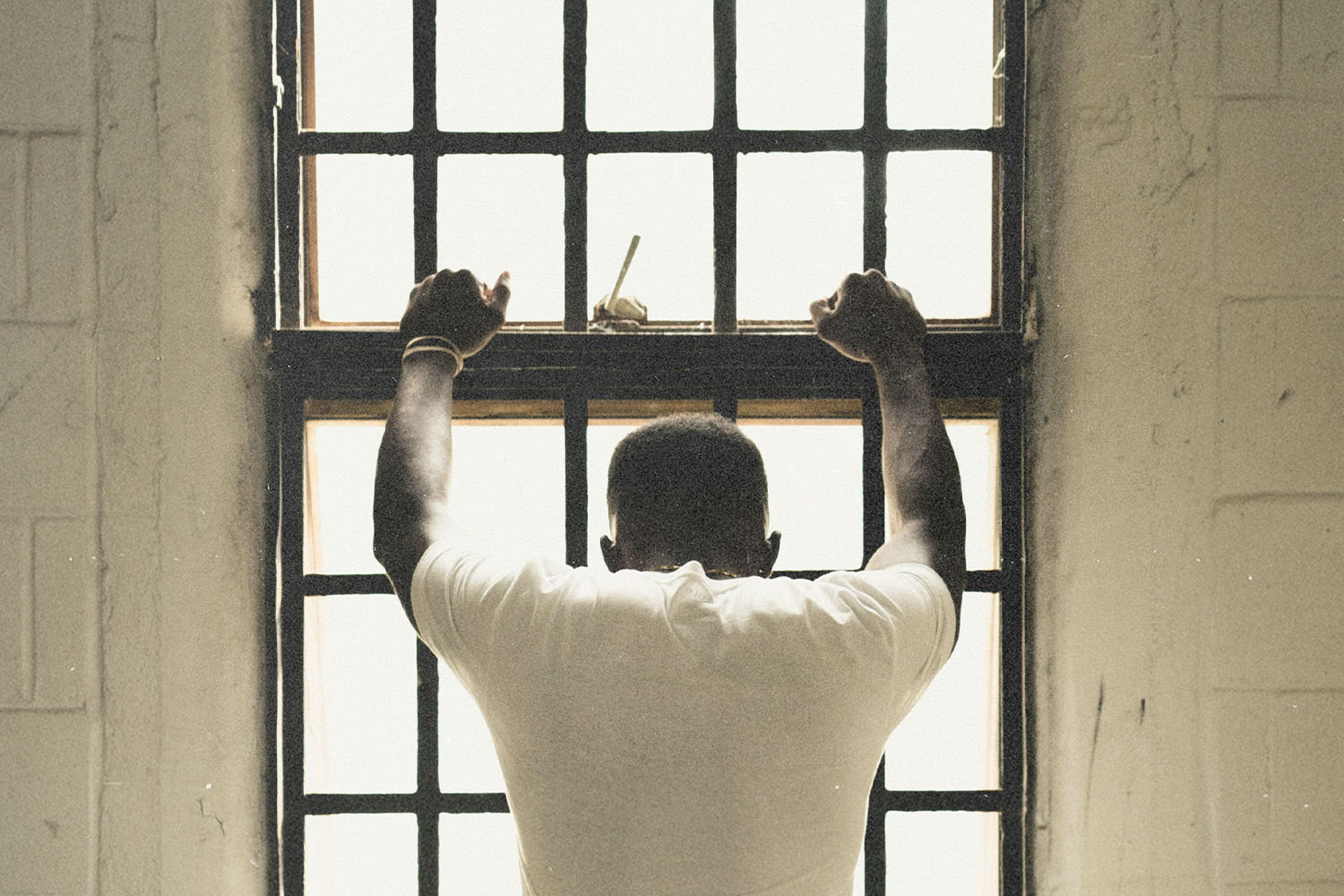 Person standing with hands gripping window bars while looking out through a bright, backlit window