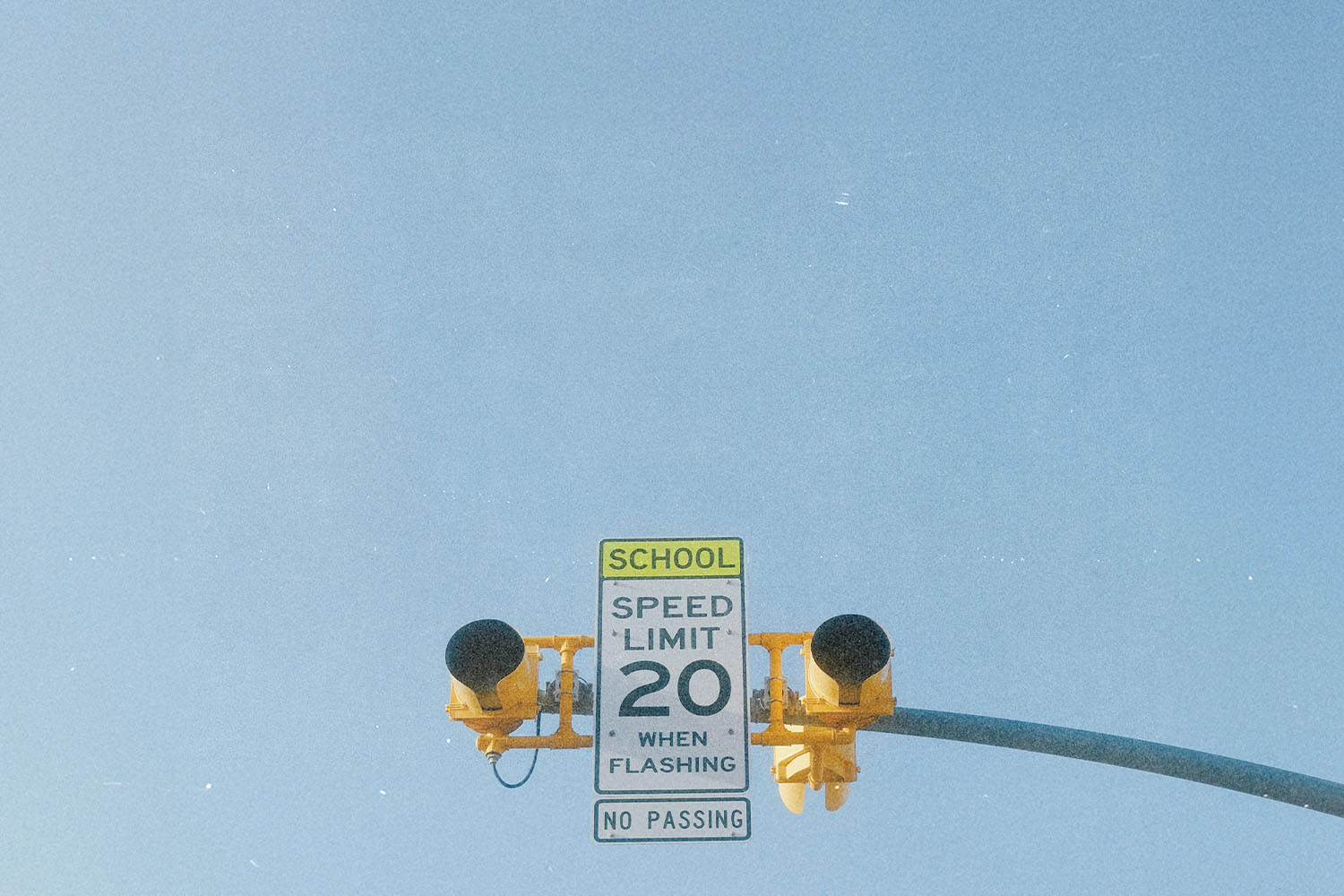 School zone speed‑limit sign reading “20 when flashing,” mounted beneath yellow traffic lights against a clear blue sky
