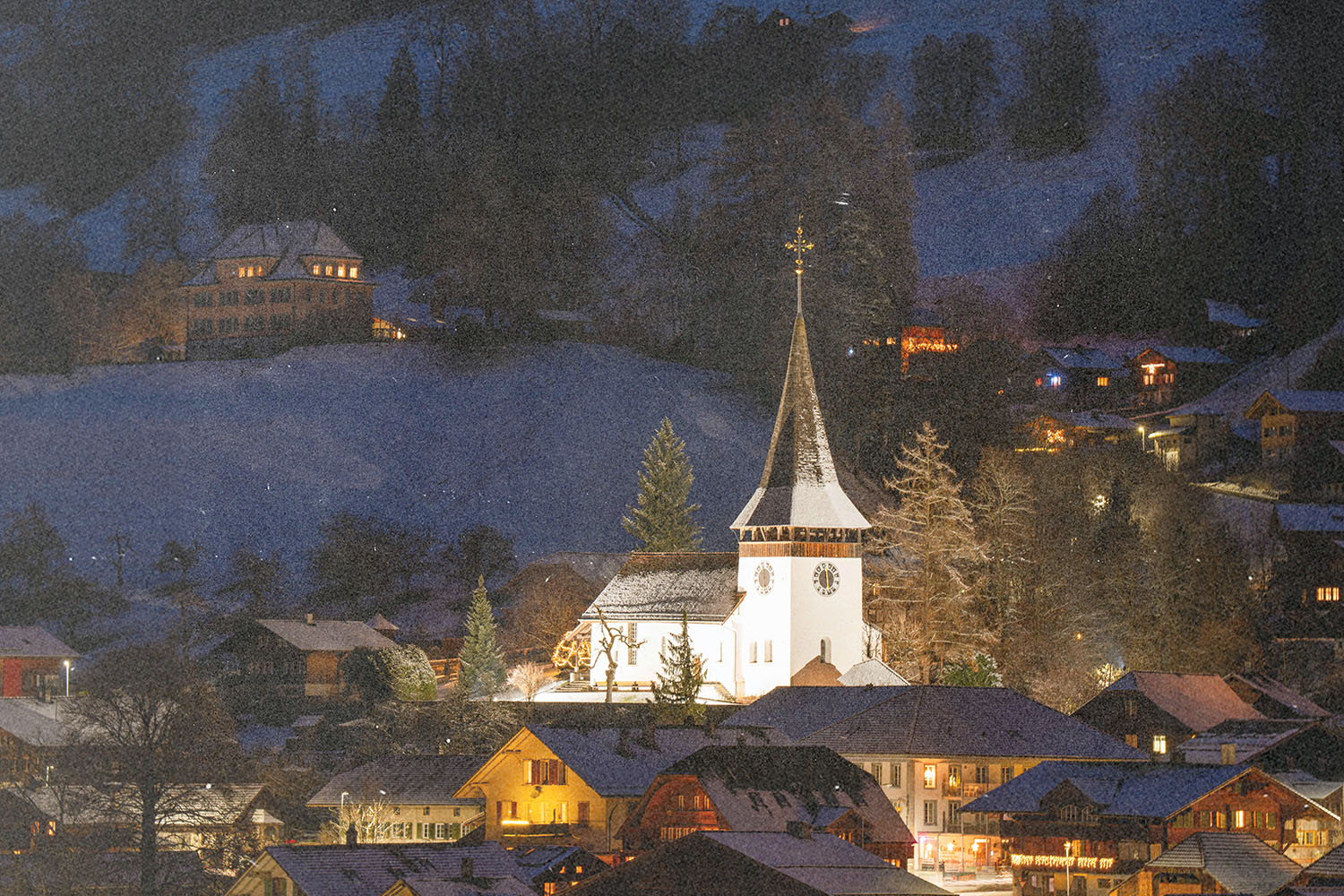 Lit church steeple surrounded by snow-covered rooftops and trees in a winter mountain village at dusk