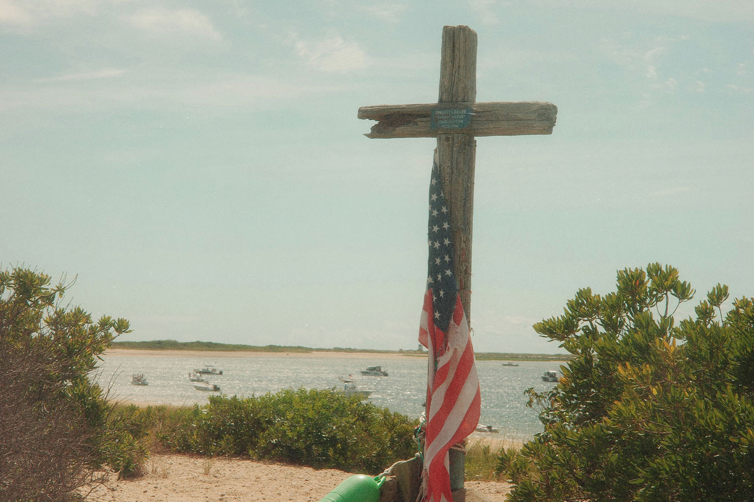 Weathered wooden cross standing on a sandy shoreline with an American flag draped over it, overlooking calm water and distant boats