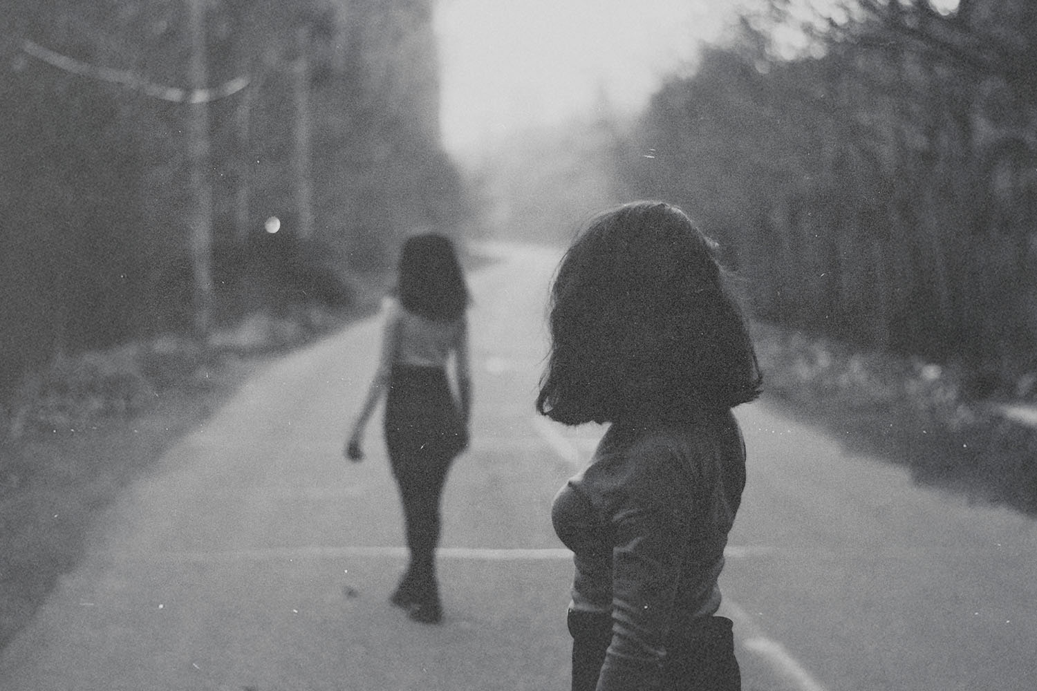 Two people walking on a quiet road lined with trees, one closer to the camera and one farther ahead
