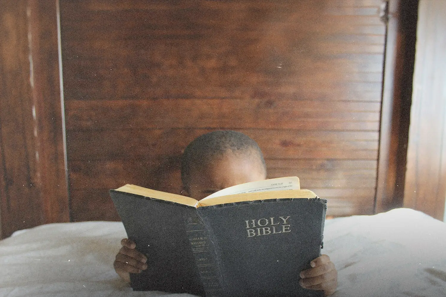 Young child holding and reading an open Bible while sitting indoors against wooden paneling