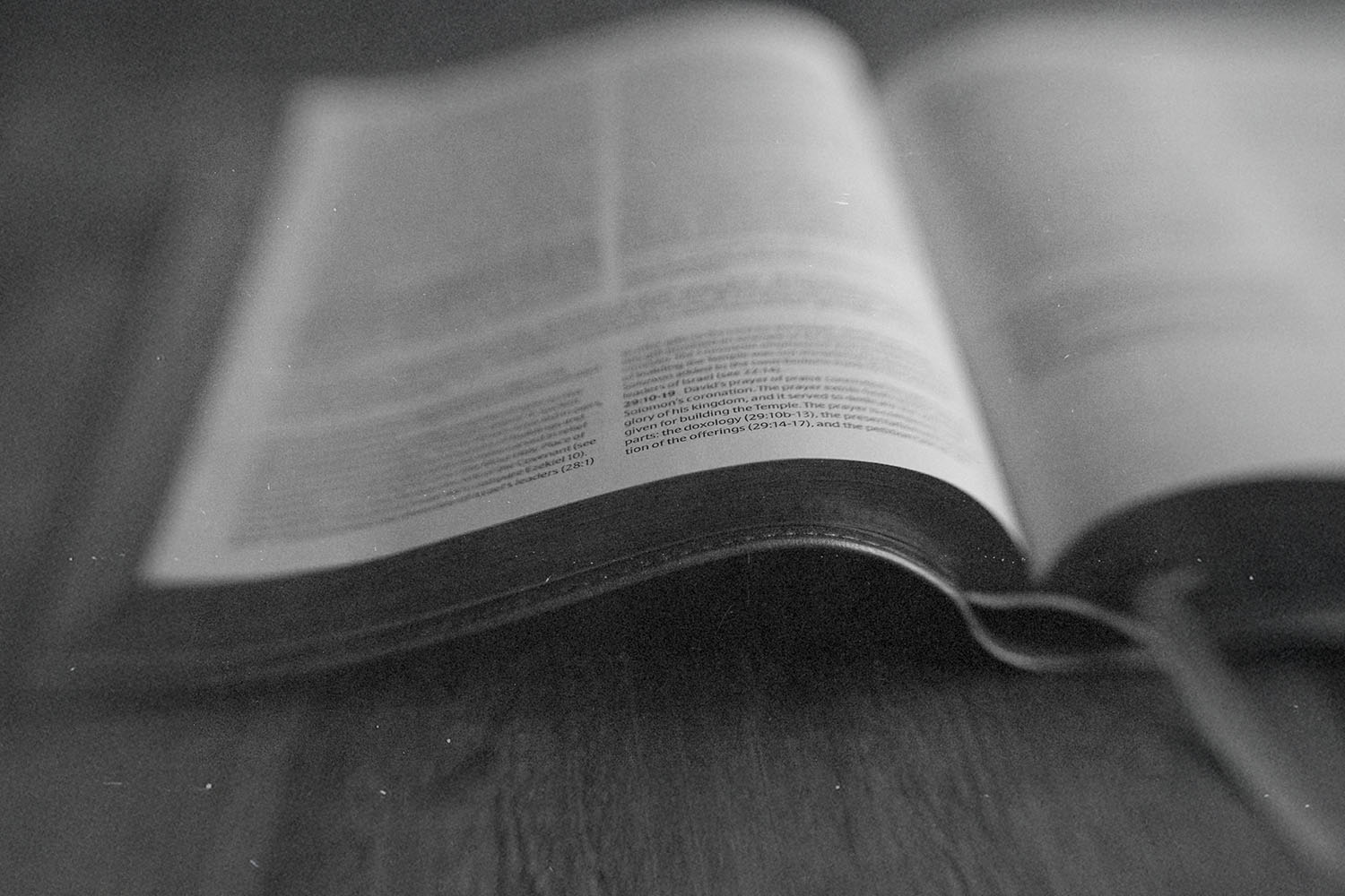 Open Bible lying on a wooden surface, with the pages softly curved and text slightly out of focus in a black‑and‑white image