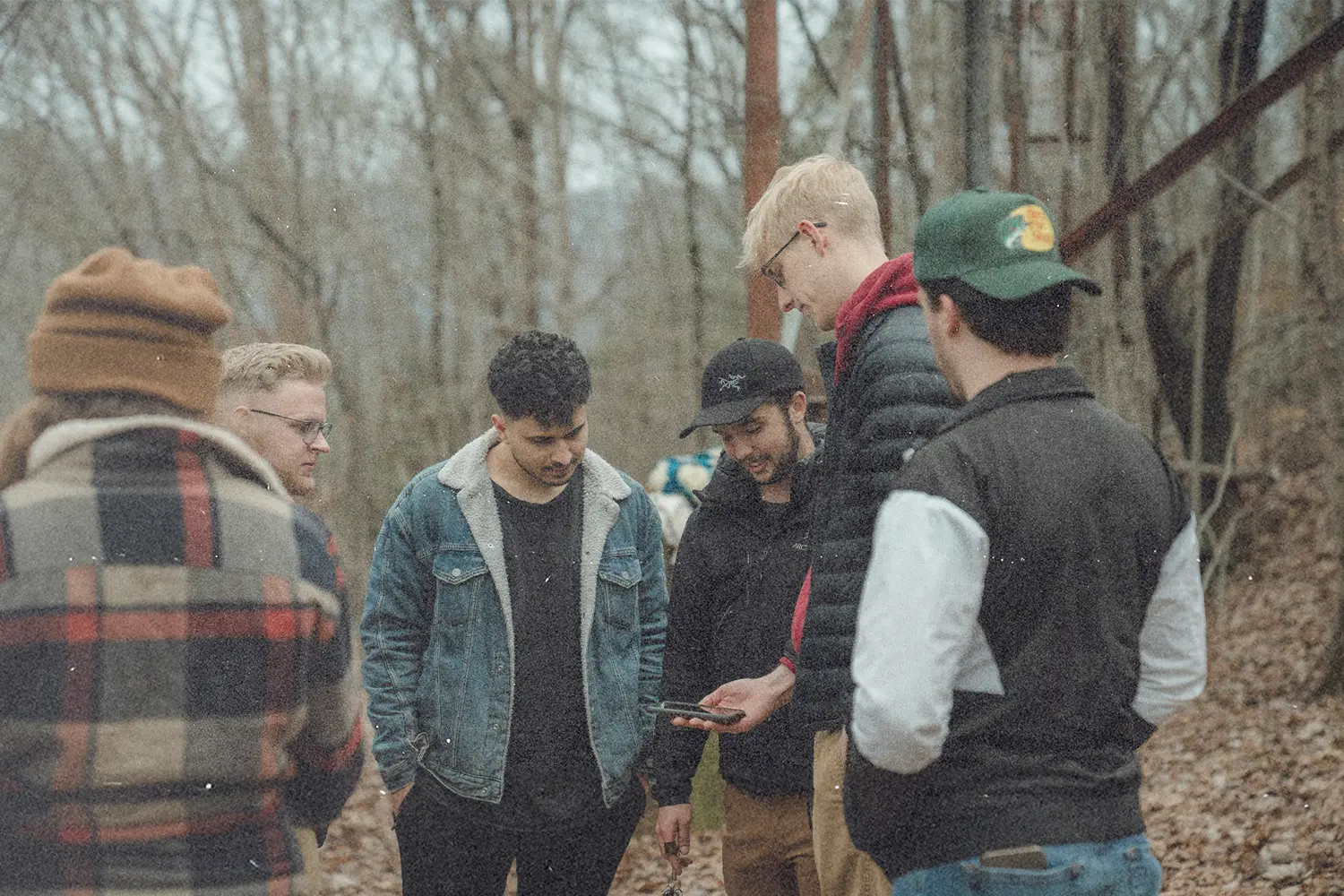 Group of people gathered in a wooded outdoor area, standing together among trees and fallen leaves