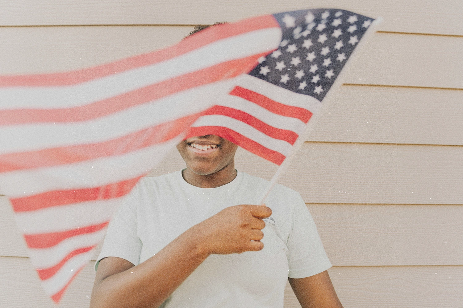 Young kid standing against a light-colored wall, waving an American flag that fills the foreground