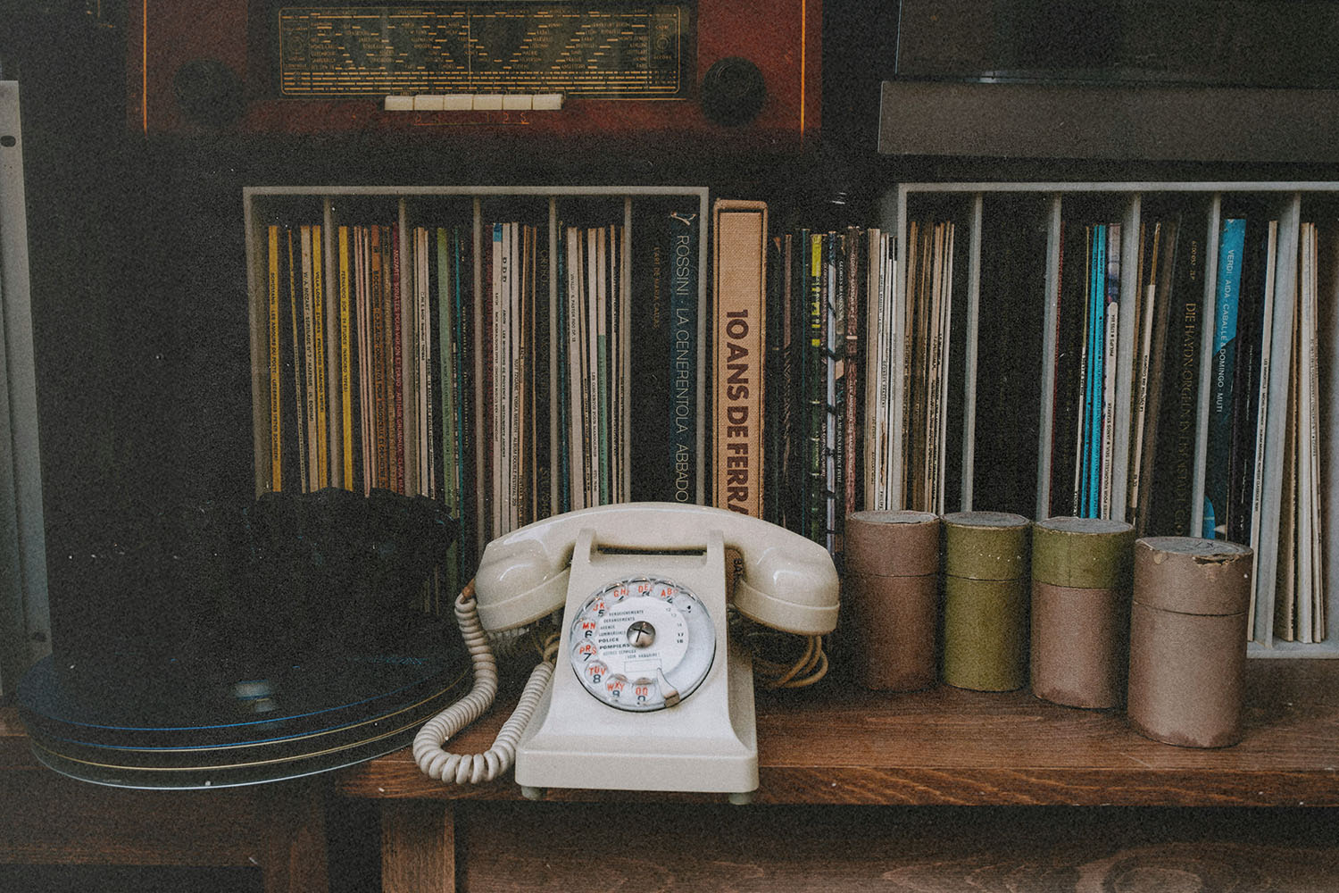 Vintage rotary telephone displayed on a wooden shelf surrounded by vinyl records, books, and small storage canisters
