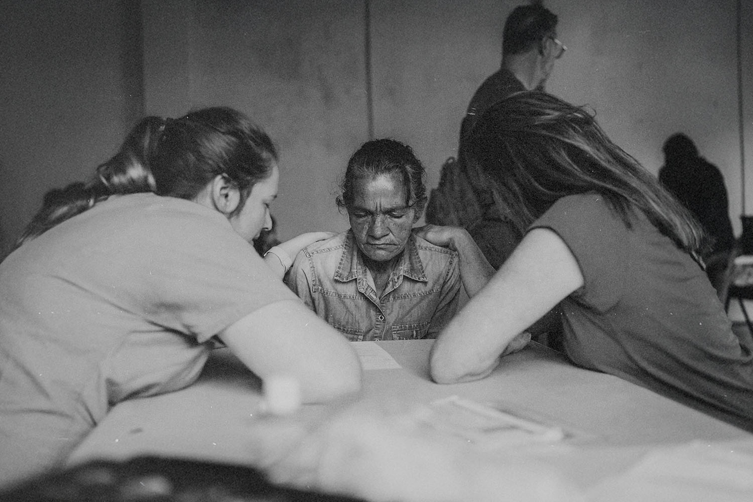 Three people sitting around a table in a quiet room, leaning in together in a moment of prayer