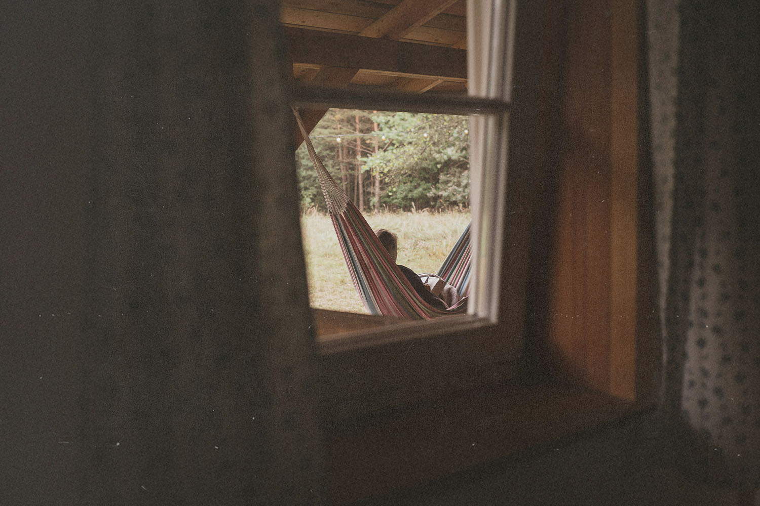 Person relaxing in a striped hammock under a wooden porch, viewed through a window looking out toward a grassy area and trees