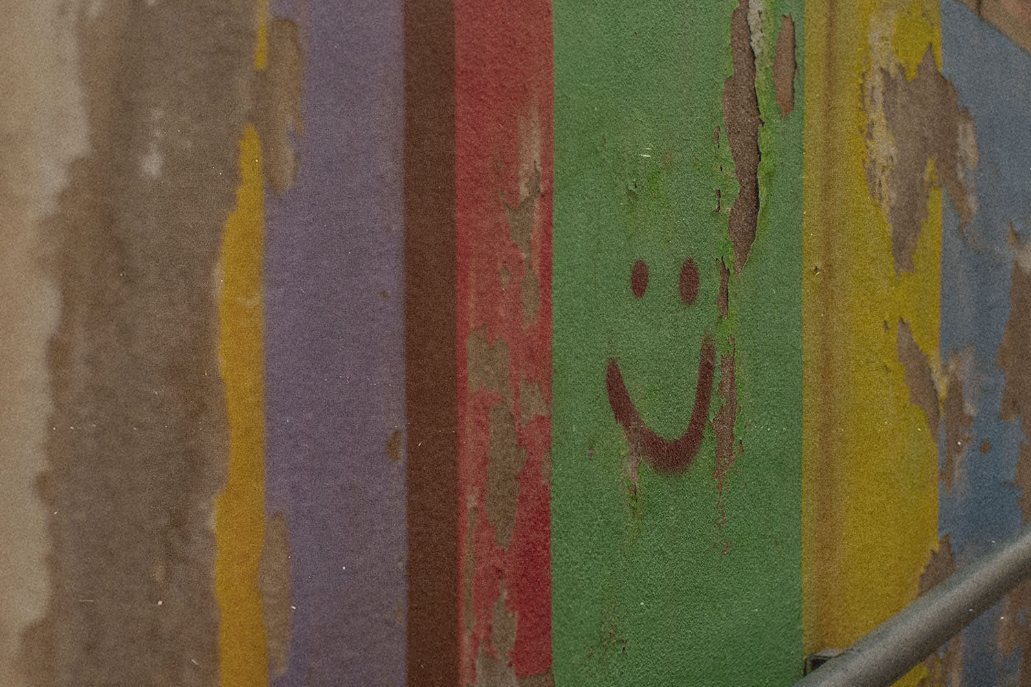 Peeling painted wall with vertical stripes of faded colors, including green, red, purple, yellow, and blue, featuring a simple smiley face drawn on the green section