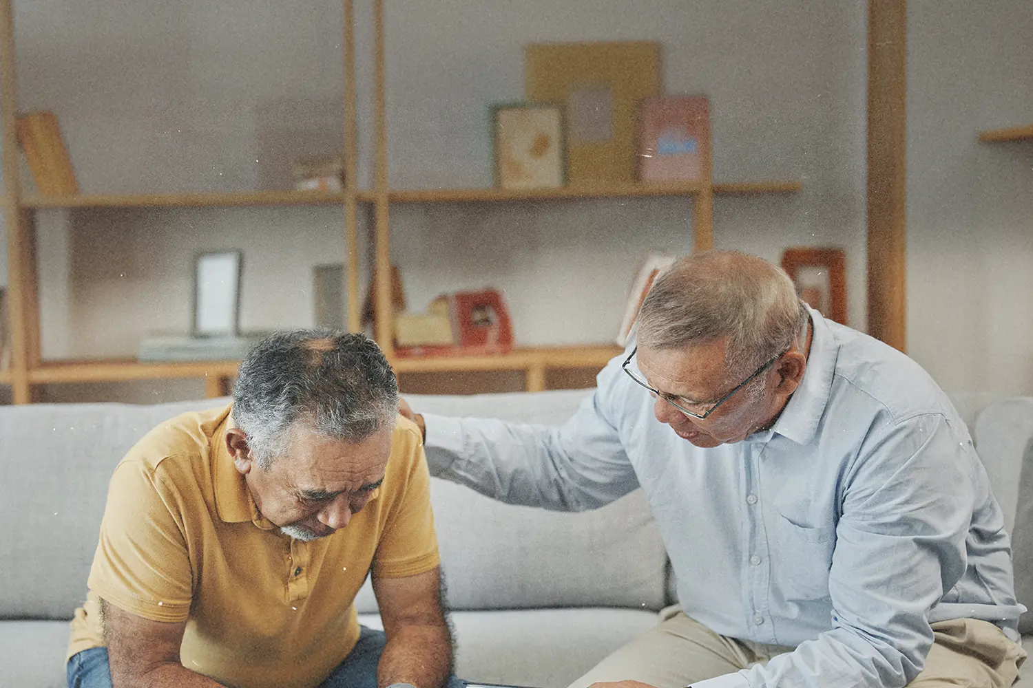 Person sitting on a couch with another person beside them offering support with a hand on their shoulder in a living‑room setting