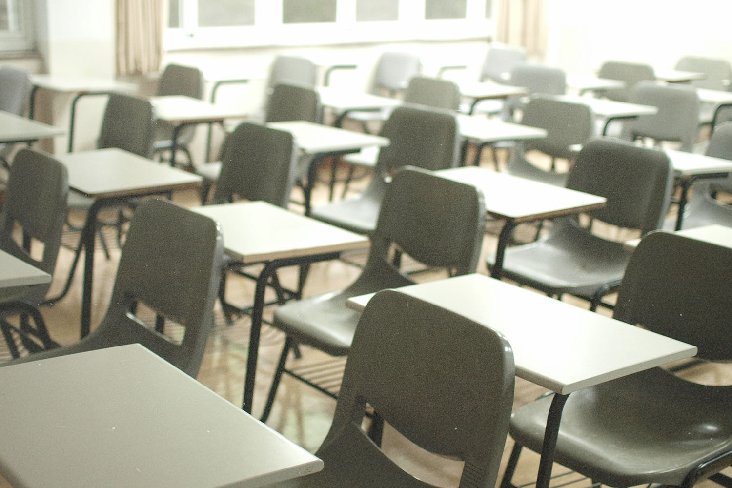 Rows of empty desks and chairs fill a bright classroom, with sunlight coming through large windows at the back of the room