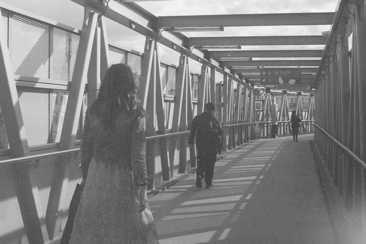 People walking through a long, covered pedestrian walkway with metal beams casting shadows along the path