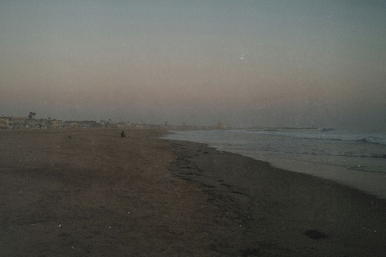 Wide, empty beach at dusk with gentle waves rolling onto the shore and faint silhouettes of distant buildings along the coastline