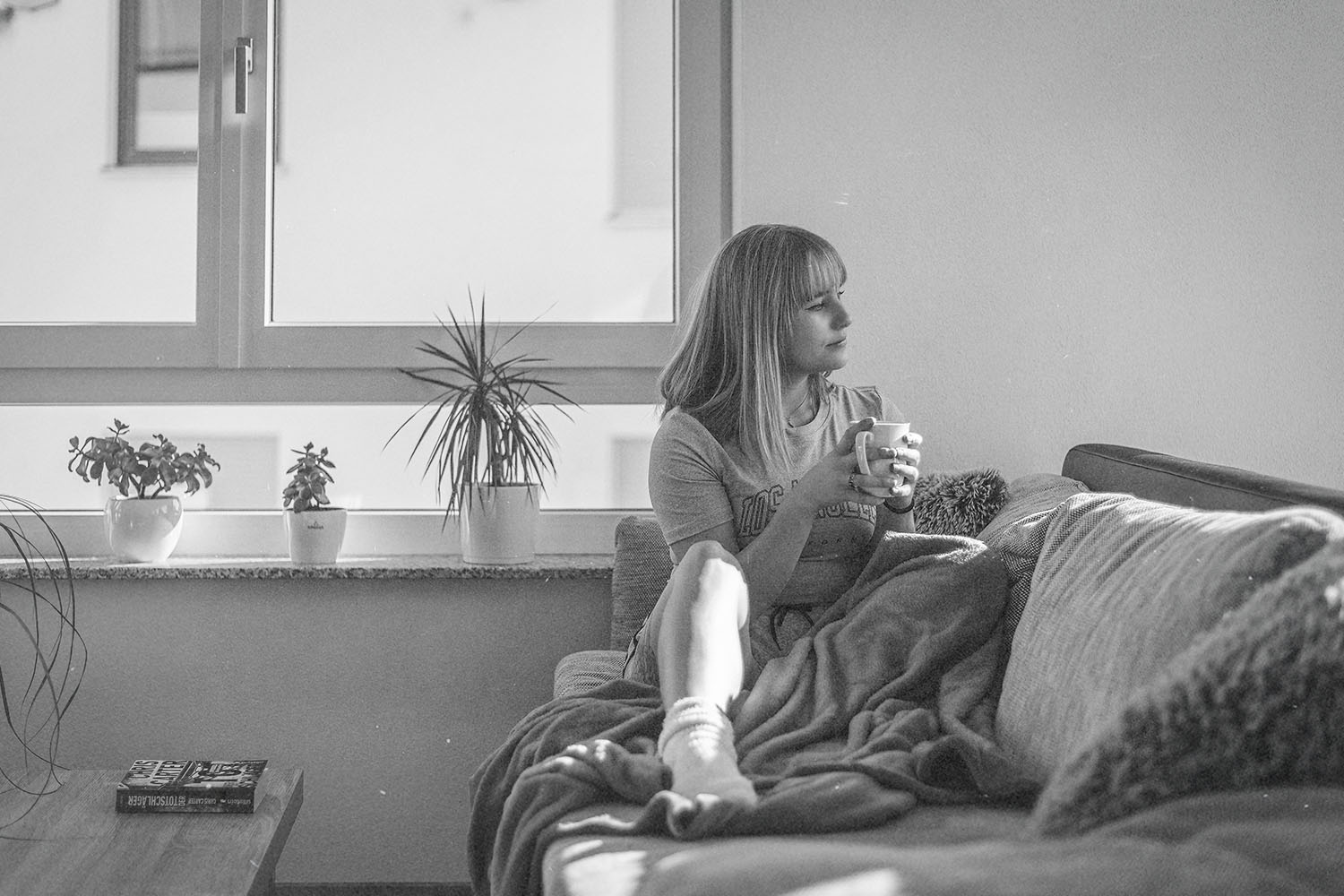 Person relaxing on a sofa with a blanket and holding a warm drink near a bright window with potted plants