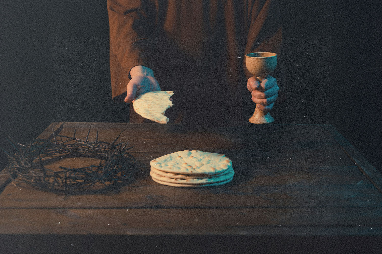 Hands holding unleavened bread and a communion cup above a wooden table with matzo and a crown of thorns