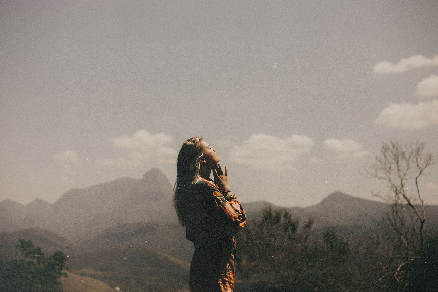 Person standing outdoors on a sunny day, holding a phone while overlooking distant mountains and a hazy landscape