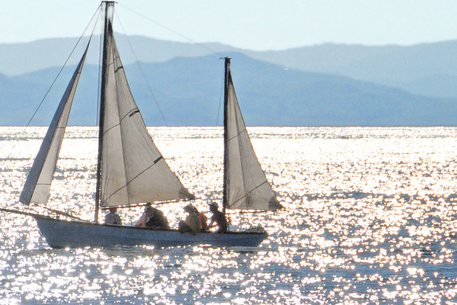 Sailboat carrying several people gliding across a sunlit lake with sparkling water and distant mountains in the background