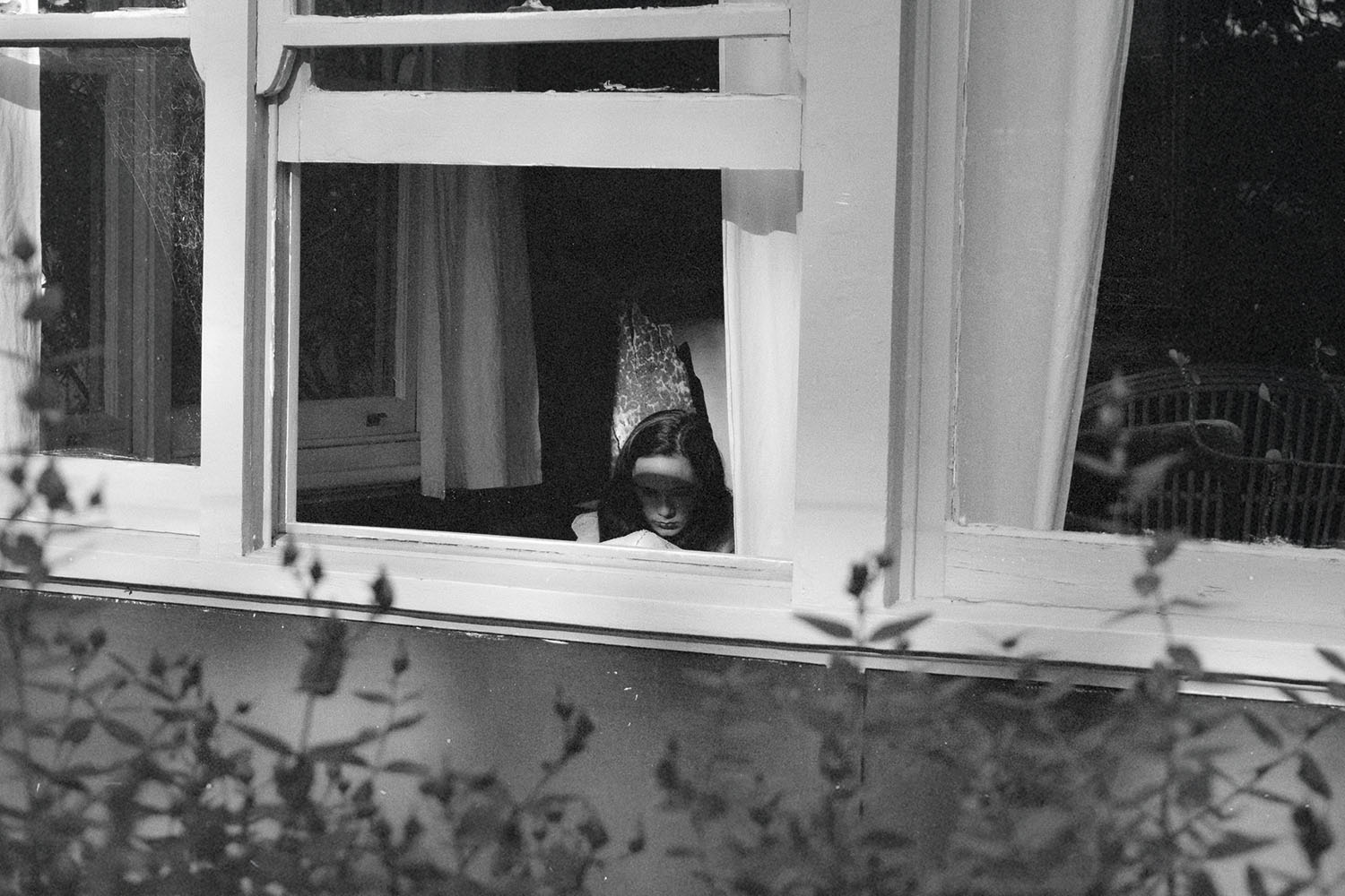 View through an open window into a dimly lit room where a person is seated near a chair, framed by white curtains and surrounding foliage