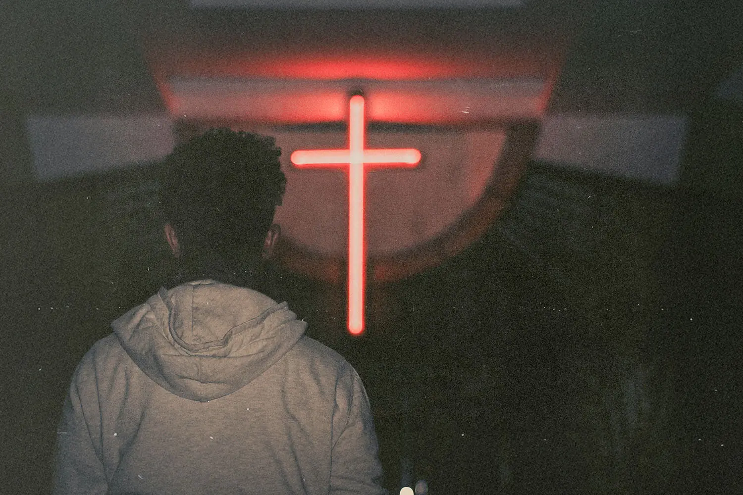 Silhouetted person standing in a dark room facing a glowing red cross on the wall