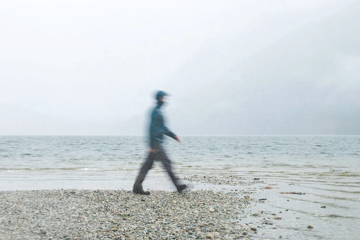 Person walking along a rocky lakeshore on a foggy day with mountains fading into the mist