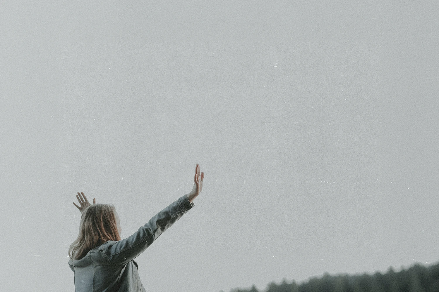 Person standing outdoors with arms raised toward the sky against a wide, soft gray backdrop and distant tree line