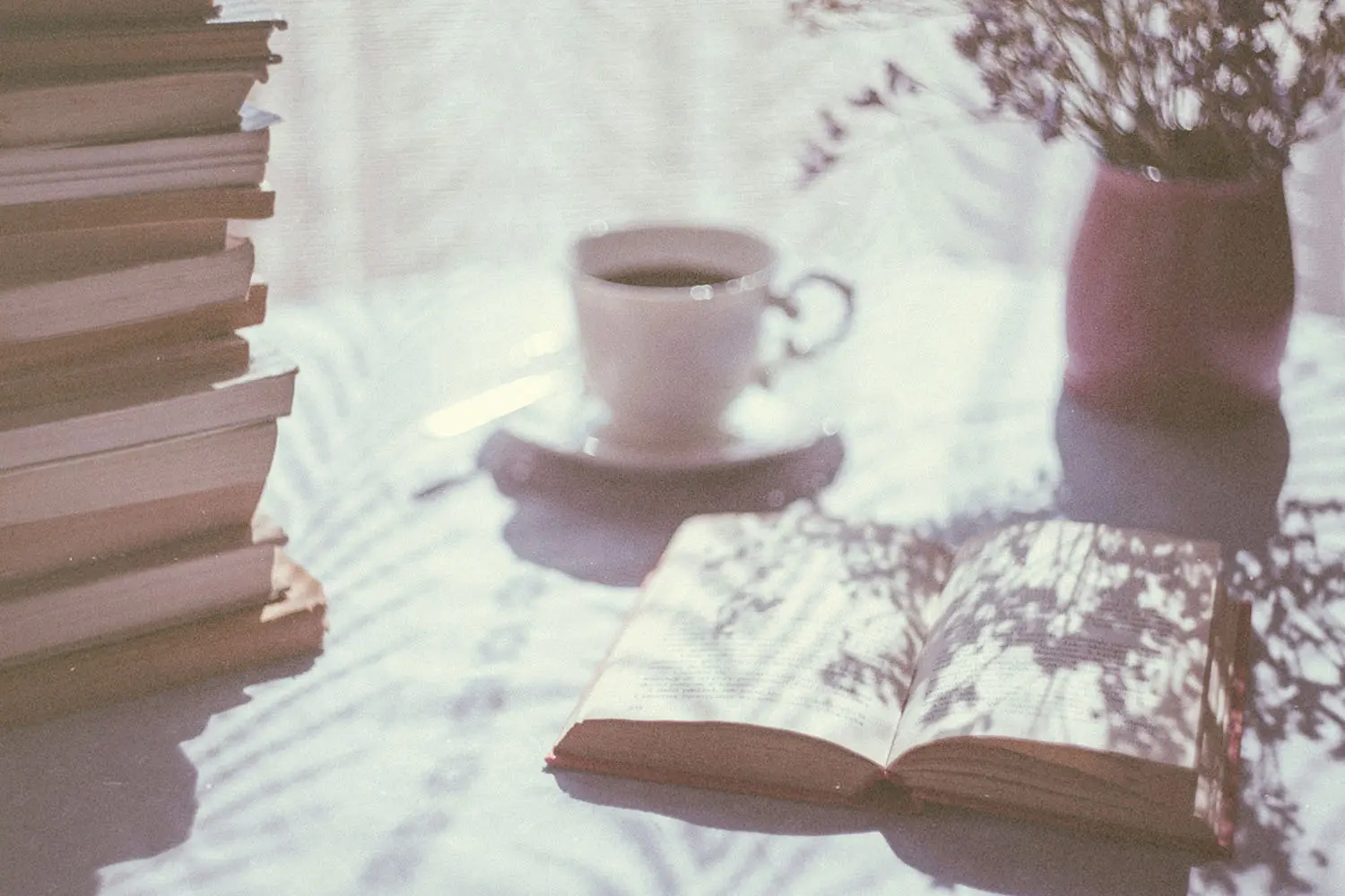 An open book rests on a sunlit table beside a tall stack of books, a cup of coffee, and a vase of flowers, with soft shadows cast across the scene