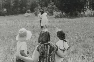 A woman stands in a grassy field with two young children at her sides, all facing toward another pair of people walking ahead across the open meadow, with trees lining the distance