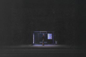 Single person stands under the dim blue light of a bus stop shelter at night, surrounded by darkness