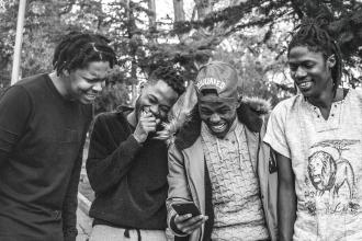 Four people stand outdoors closely together, looking at a smartphone one person is holding
