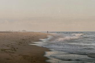 A lone person walks along a quiet shoreline as gentle waves roll onto the sand in the early morning light