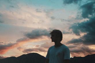 A person stands outdoors at sunset with mountains silhouetted behind them and colorful clouds filling the sky