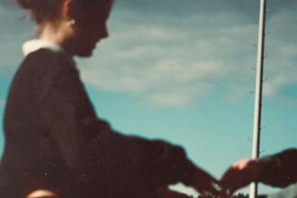 A softly focused outdoor scene showing a person in a dark long‑sleeve top holding hands with another person. A tall flagpole rises into a partly cloudy blue sky in the background.