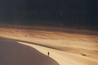 A solitary person stands on a sunlit sand dune, surrounded by vast, sweeping desert terrain under a darkened sky