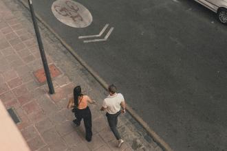 Two people walk side by side on a city sidewalk, viewed from above as they move along the street