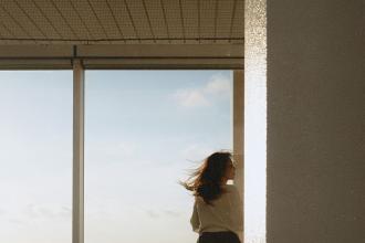 Silhouetted person near a window frame, gazing outdoors at a clear sky with soft daylight