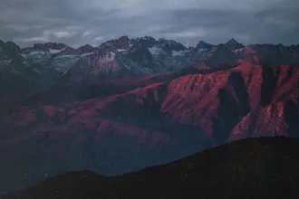 Mountain range at sunset with rugged peaks and slopes glowing red under dramatic clouds