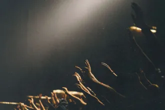 Several raised hands reaching upward toward a bright spotlight in a dark, crowded setting