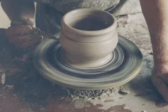 Hands shaping a clay pot on a spinning pottery wheel, with tools and wet clay visible in the workspace