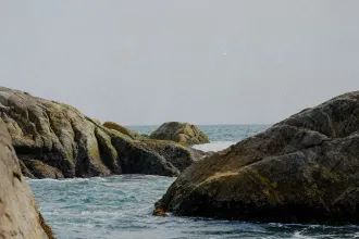 Rocky shoreline with ocean waves moving between large weathered boulders under a pale, hazy sky