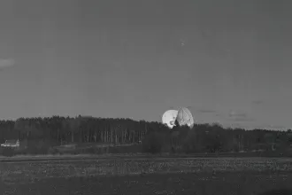 Large satellite dishes rising above a dense tree line in a wide, open field under a dark sky