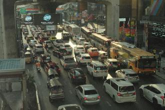 Heavy traffic jam on a multi‑lane city street filled with cars, buses, and tuk‑tuks beneath an elevated roadway in a busy urban shopping district