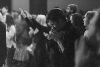 Group of people standing closely together in a dimly lit room with several individuals raising their hands and one person in the foreground holding their hands together in front of their face