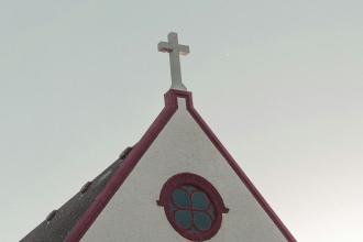 Steeple of a small church with a white cross on top and a circular stained‑glass window, set against a pale sky