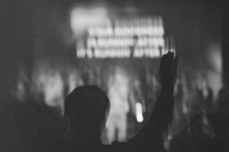 Silhouetted person raising one hand in front of a brightly lit stage with indistinct figures and text in the background