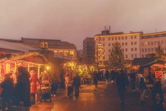 Crowds walking through an outdoor Christmas market at dusk, with warm lights glowing from decorated stalls and buildings in the background