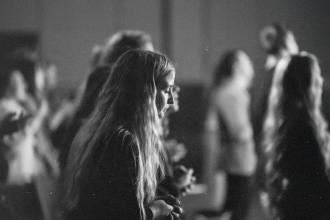 People standing in a dimly lit room during what appears to be a worship or group gathering, with individuals facing forward in soft focus