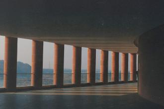 Long covered walkway with tall red columns casting angled shadows across the floor, overlooking a distant landscape in soft morning light