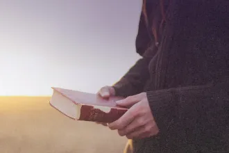 A person stands outdoors at sunrise or sunset, holding an open, worn Bible in both hands as warm light glows across the landscape
